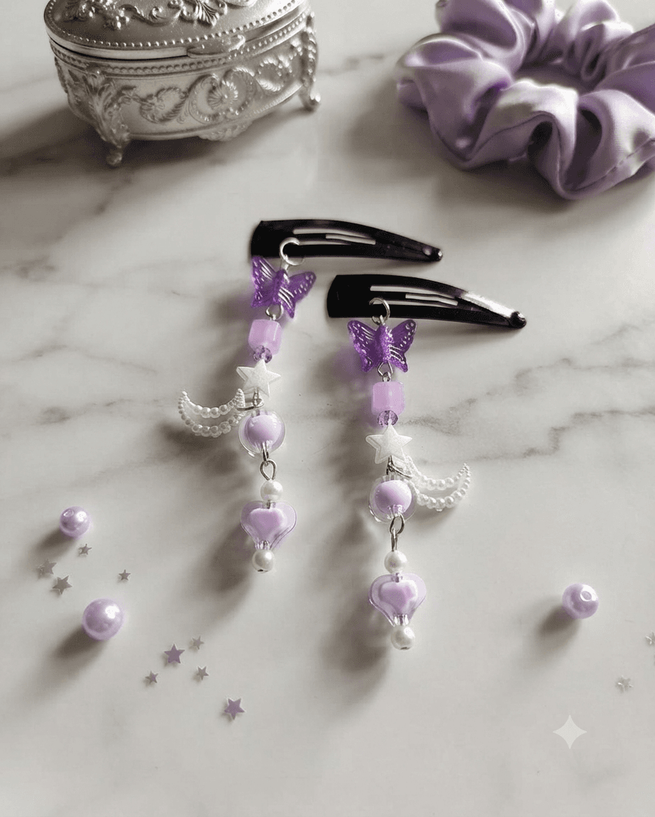 Pair of purple butterfly hair clips with lavender beads, white stars, crescent charms and heart details displayed on a marble-style surface.
