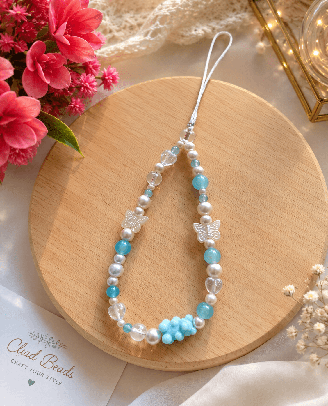 Aqua blue, white and clear beaded phone strap displayed on a round wooden board with butterfly and heart-shaped beads.