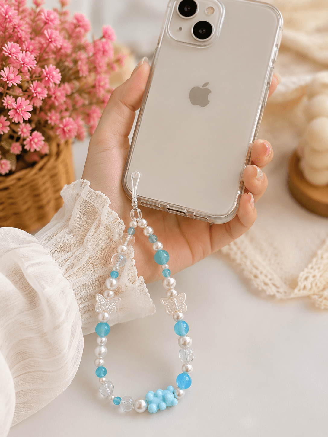 Aqua blue handmade beaded phone strap attached to a clear phone case and held in hand, featuring butterfly, heart and pearl-look bead details.