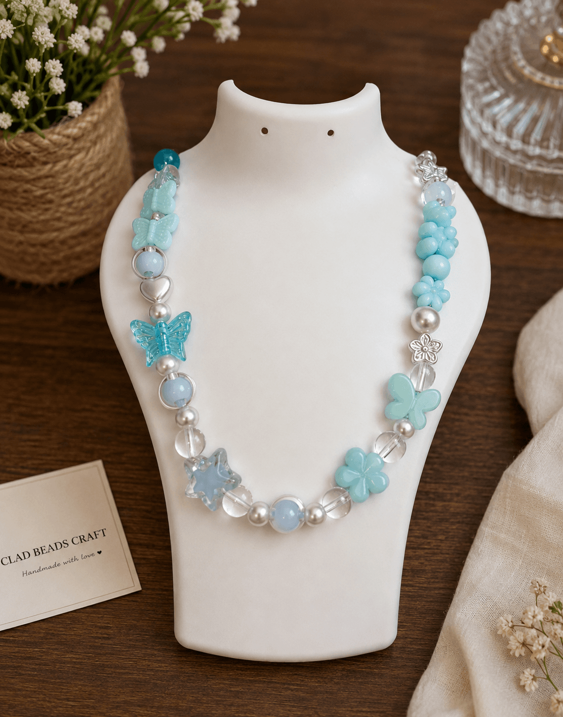 Aqua blue handmade beaded necklace with butterfly, flower, clear and white beads displayed on a white jewellery bust.