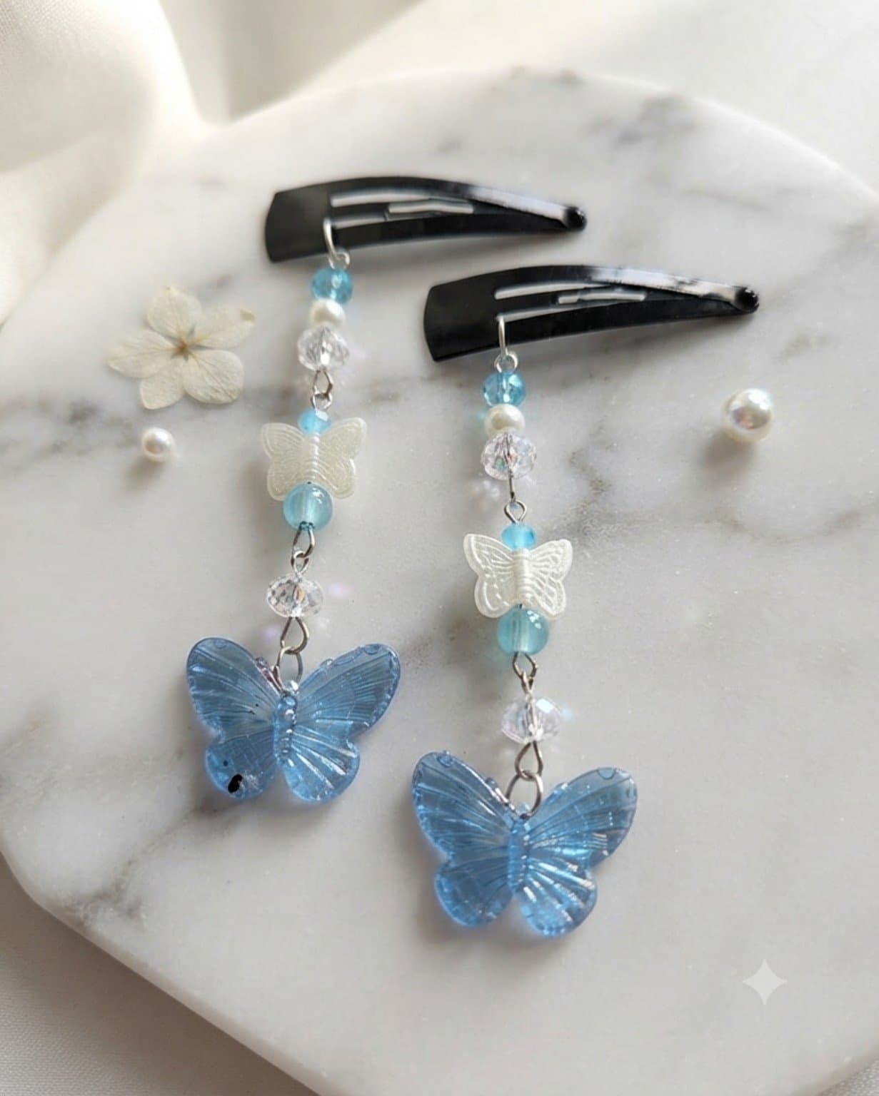 Pair of handmade blue butterfly dangle hair clips with white butterfly details, blue beads and clear faceted beads displayed on a marble-style surface