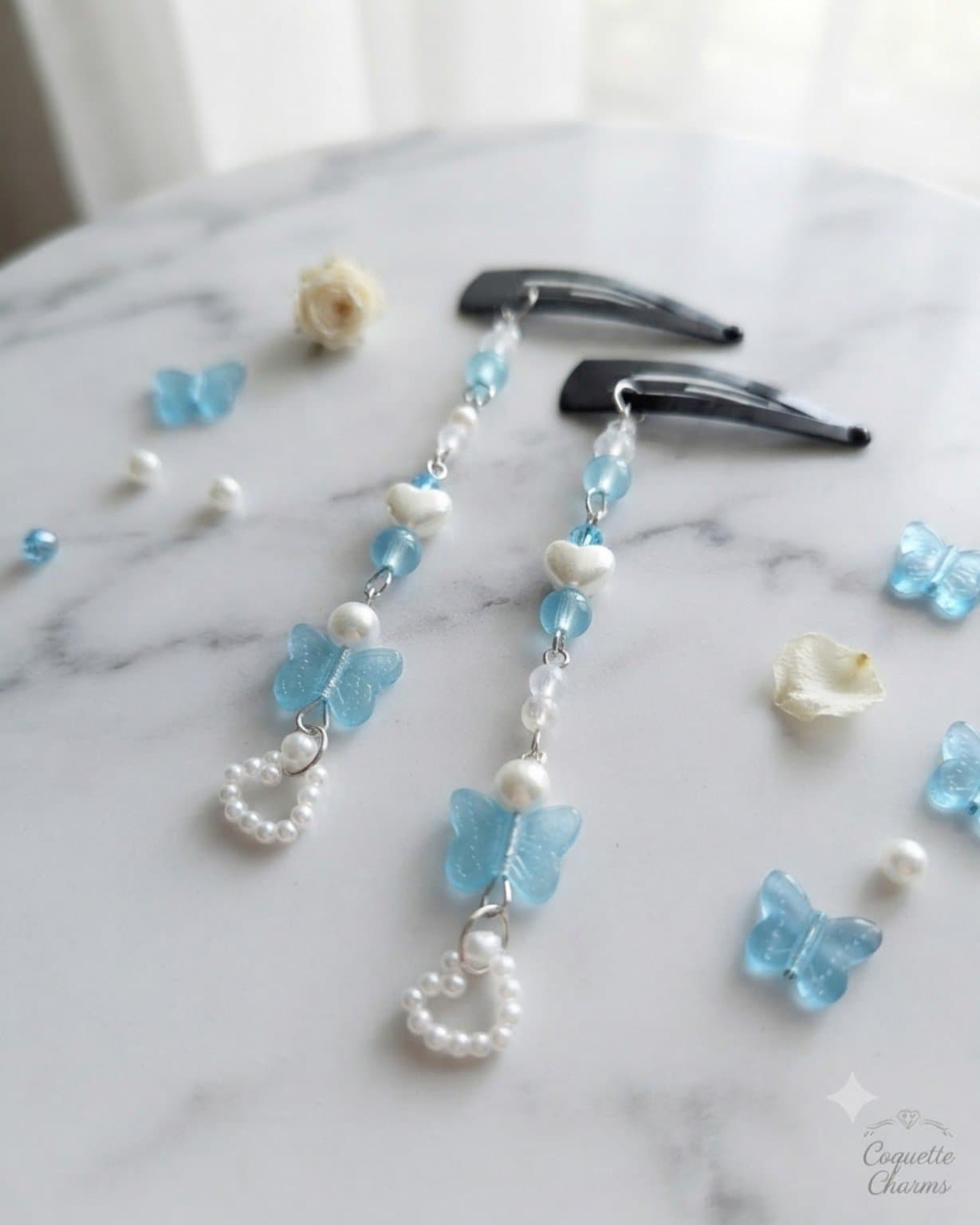 Pair of handmade aqua blue butterfly hair clips with white heart beads and dangling heart charms displayed on a marble-style surface.