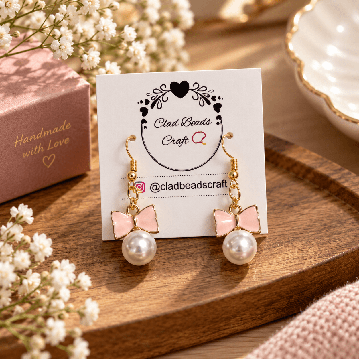 Pair of handmade pink bow drop earrings with white acrylic pearl beads and gold-plated hooks displayed on a Clad Beads Craft card.