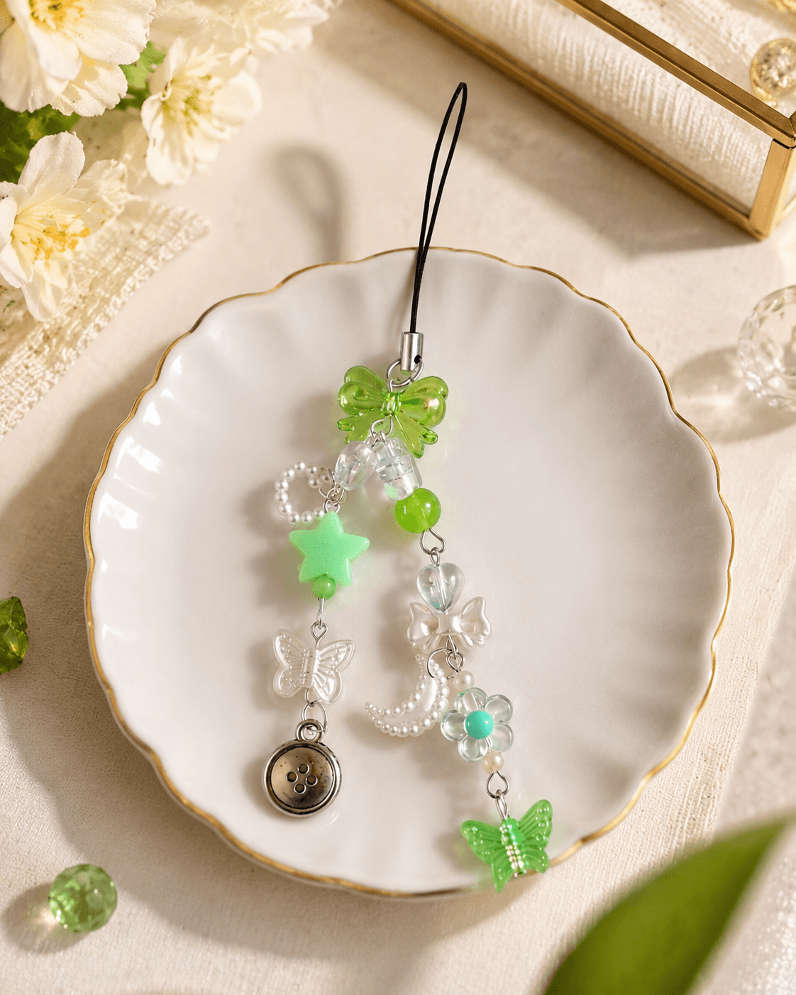 Handmade green beaded phone charm strap with a translucent green bow, green star, clear butterfly, floral detail, pearl-look accents and round button charm displayed on a white plate.
