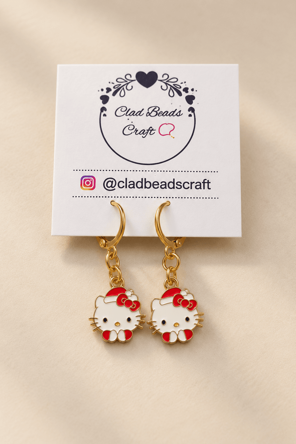 : Pair of Hello Kitty-inspired red and white character charm earrings with gold-plated hoops displayed on Clad Beads Craft card