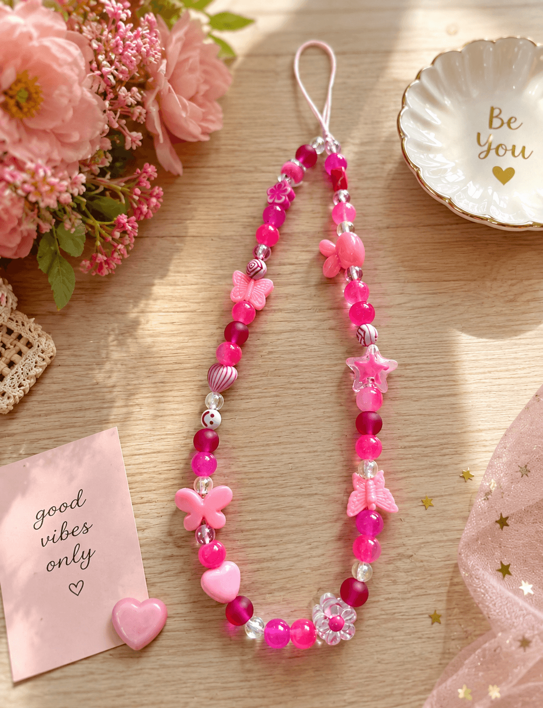 Pink and clear handmade beaded phone strap displayed on a wooden surface with butterfly, flower, star and heart details.