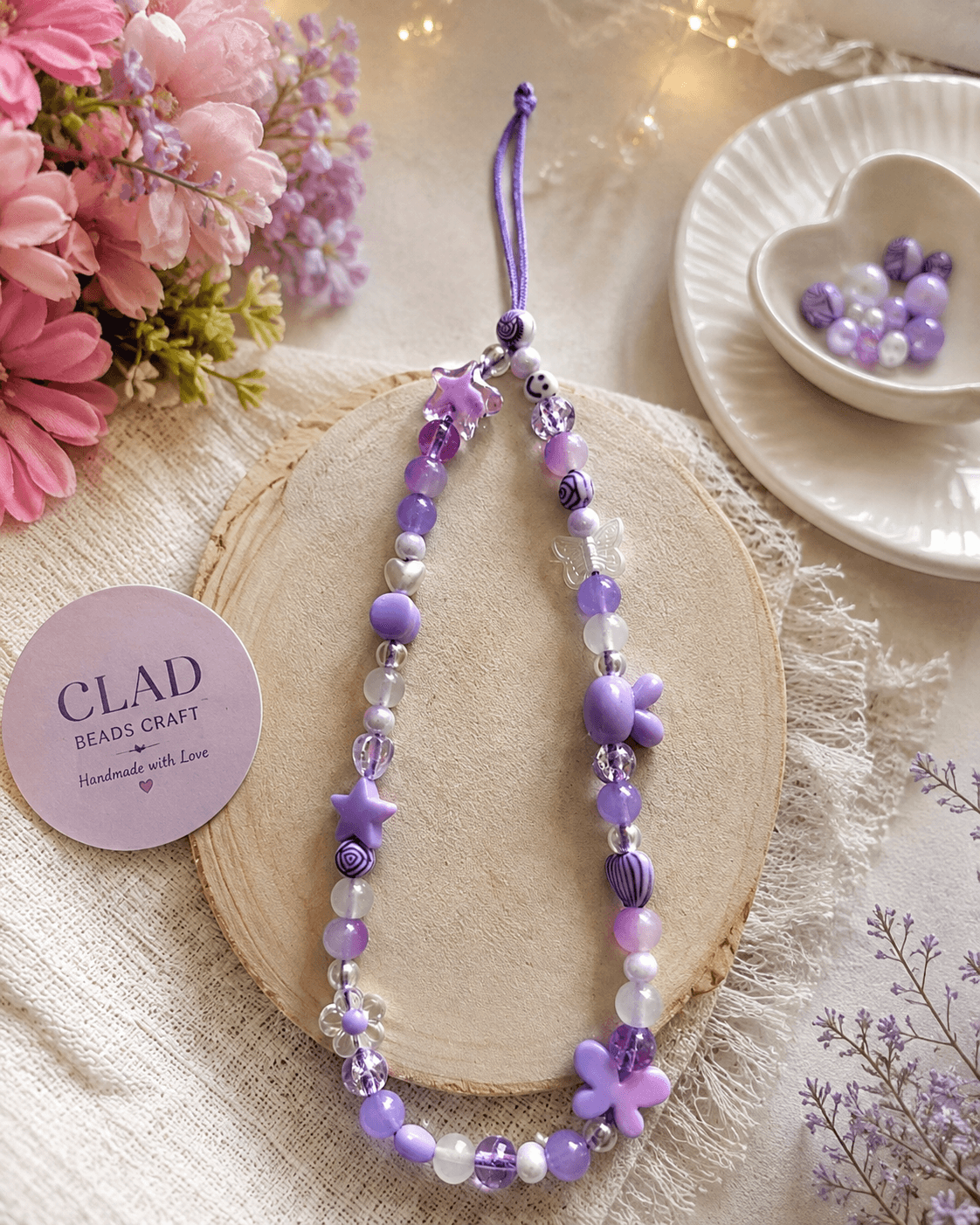 : Purple, white and clear handmade phone strap displayed on a wooden board with butterfly, teddy, flower, star and patterned beads.