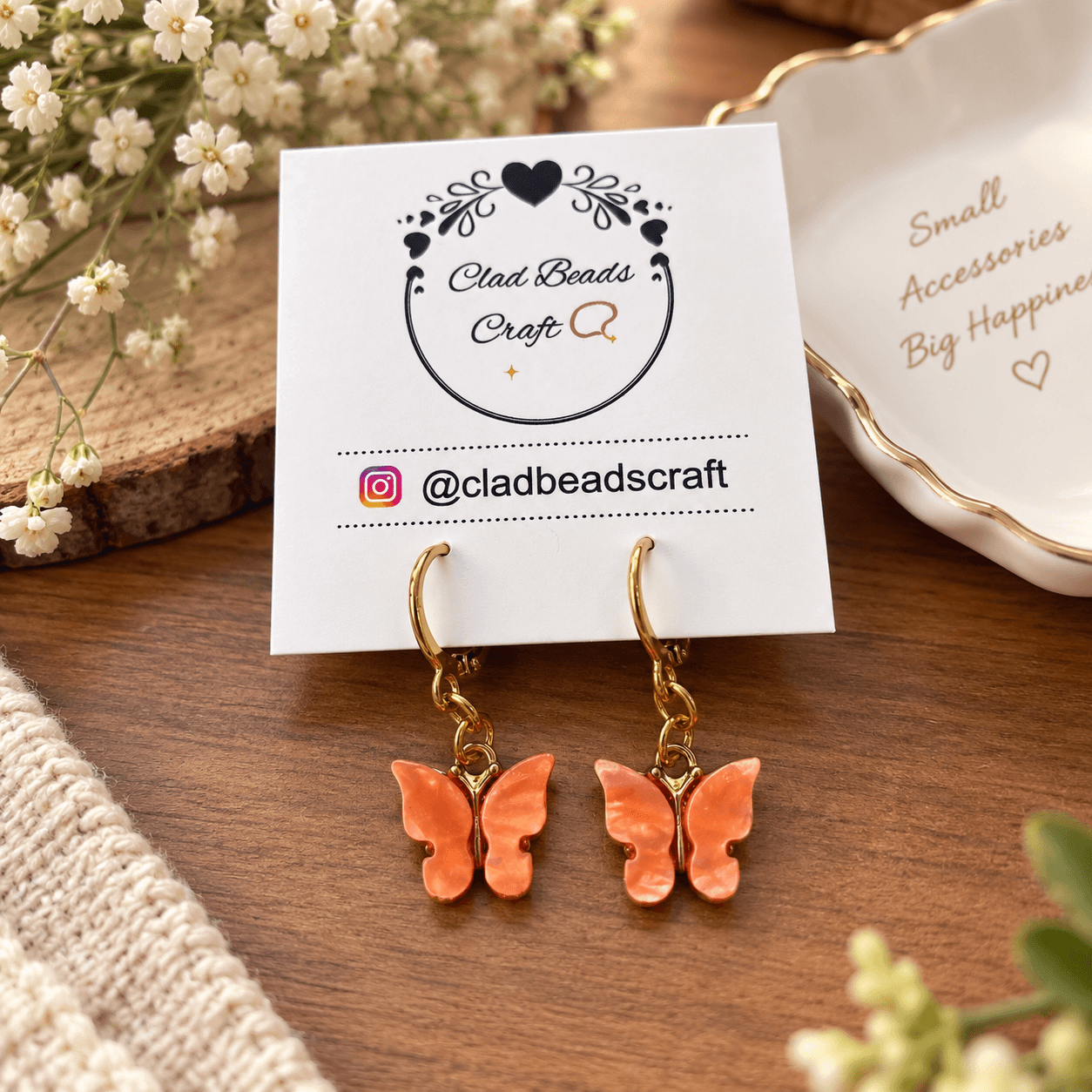 Pair of coral-orange butterfly dangle earrings with gold-tone hoops displayed on a Clad Beads Craft card