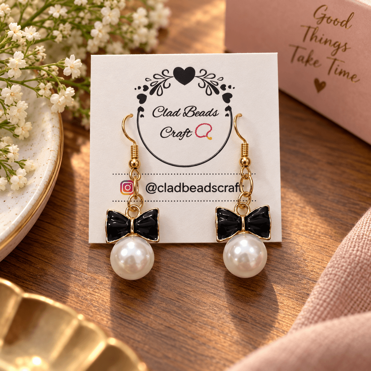 Handmade black bow drop earrings with white pearl-look beads, gold-plated hooks and gold-toned chain details displayed on a Clad Beads Craft card.