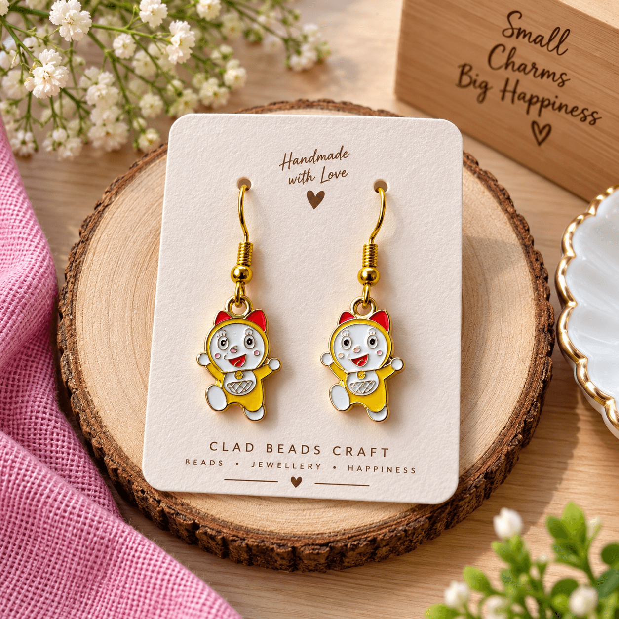 Pair of yellow cartoon charm drop earrings with red bow details and gold-tone hooks displayed on a Cladbeadscraft jewellery card.