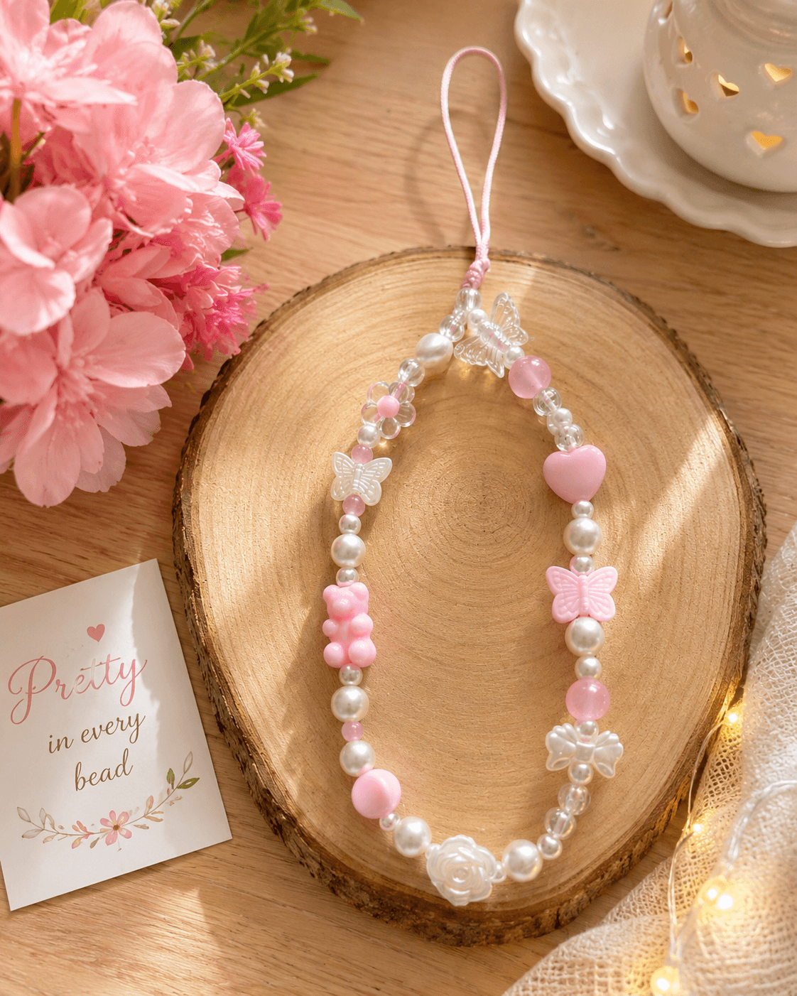Pink, white and clear handmade phone strap displayed on a wooden slice with butterfly, heart, flower and rose-inspired beads.