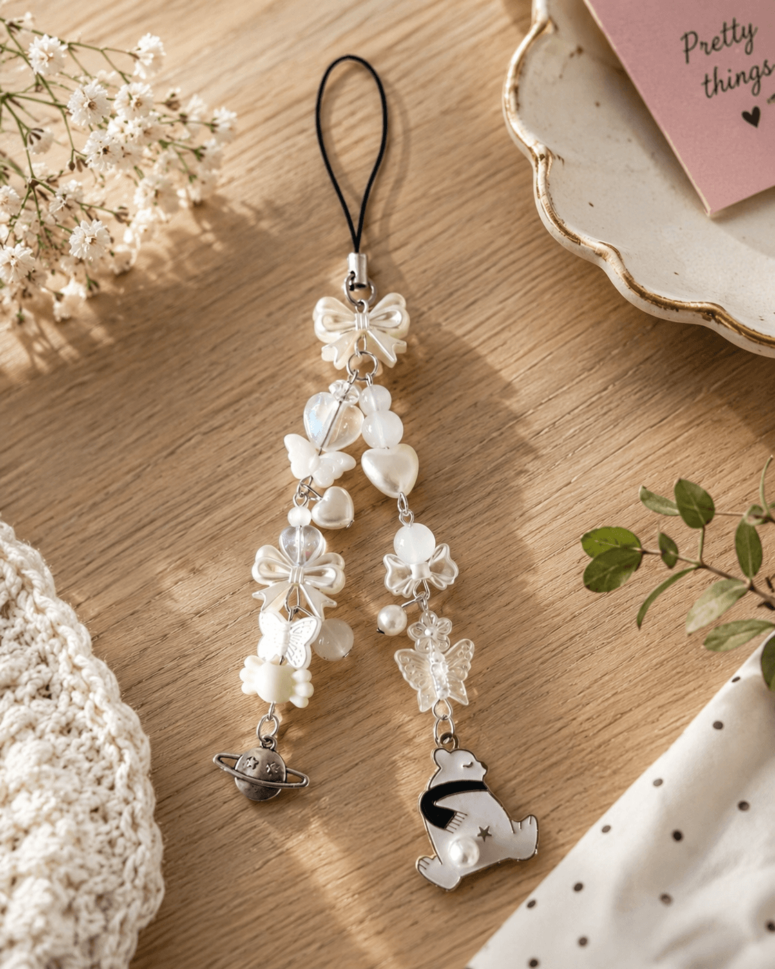 Handmade ivory and white phone charm strap with bows, butterflies, hearts, pearl-look accents, bear charm and silver-tone planet detail displayed on a wooden background.