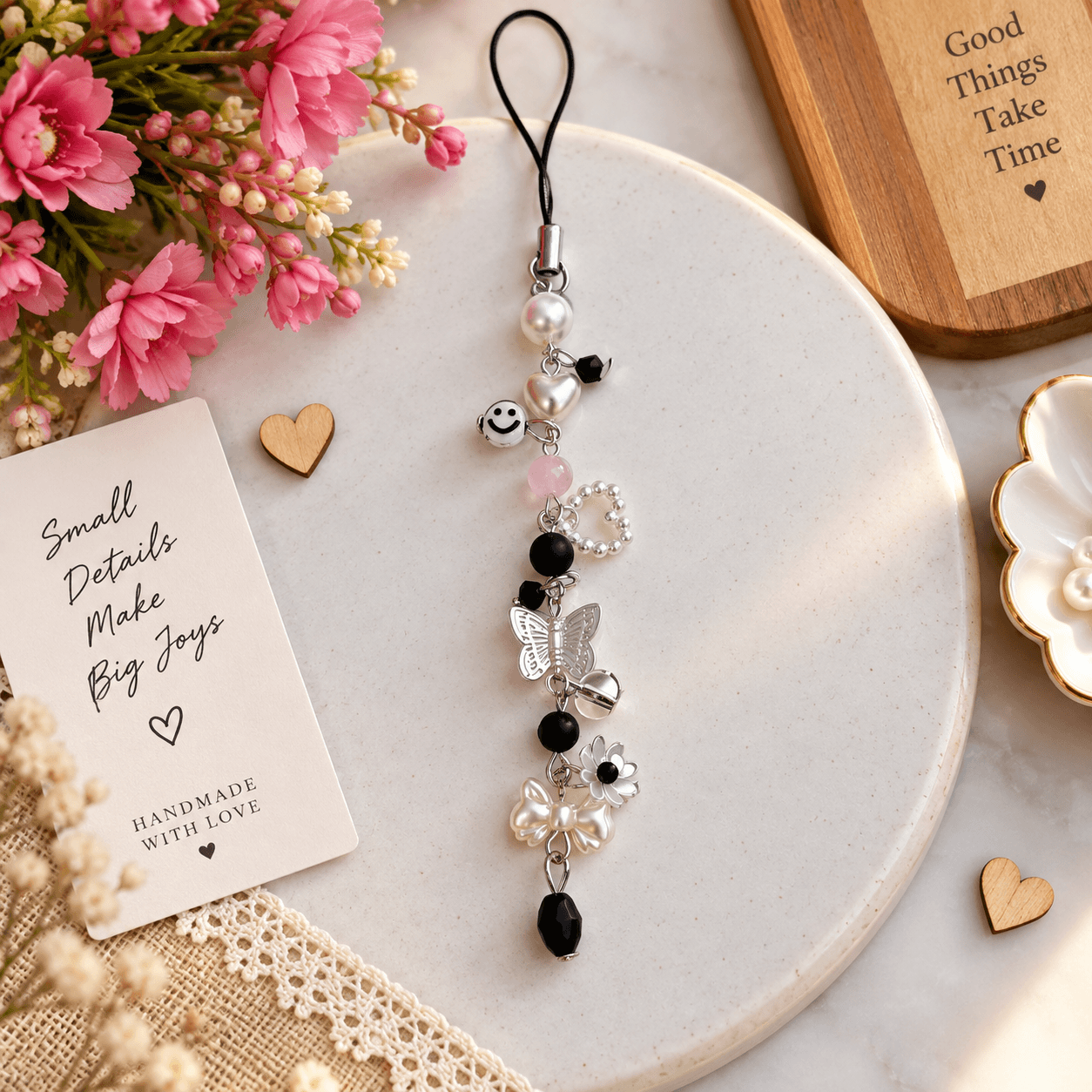 Handmade black and pink phone charm displayed on a white plate with pearl-look beads, smiley face, butterfly, flower, bow, heart and black faceted decorative bead.