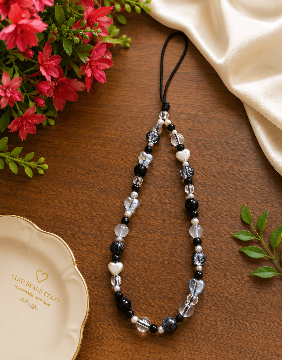 Handmade black and white beaded phone strap with clear, frosted and iridescent heart details arranged on a wooden surface.