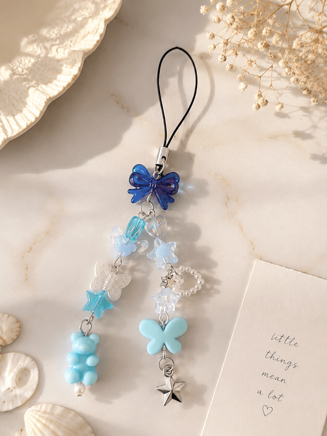 Handmade blue and pastel phone charm strap with bow, butterfly, heart, star, teddy bear, pearl-look and silver-tone details displayed on a soft background.