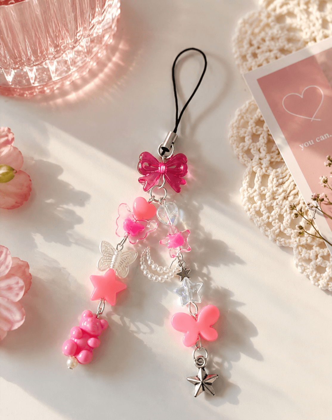 Handmade pink and clear phone charm strap with bow, heart, butterfly, flower, star, teddy bear and pearl-look details displayed on a soft pastel background.