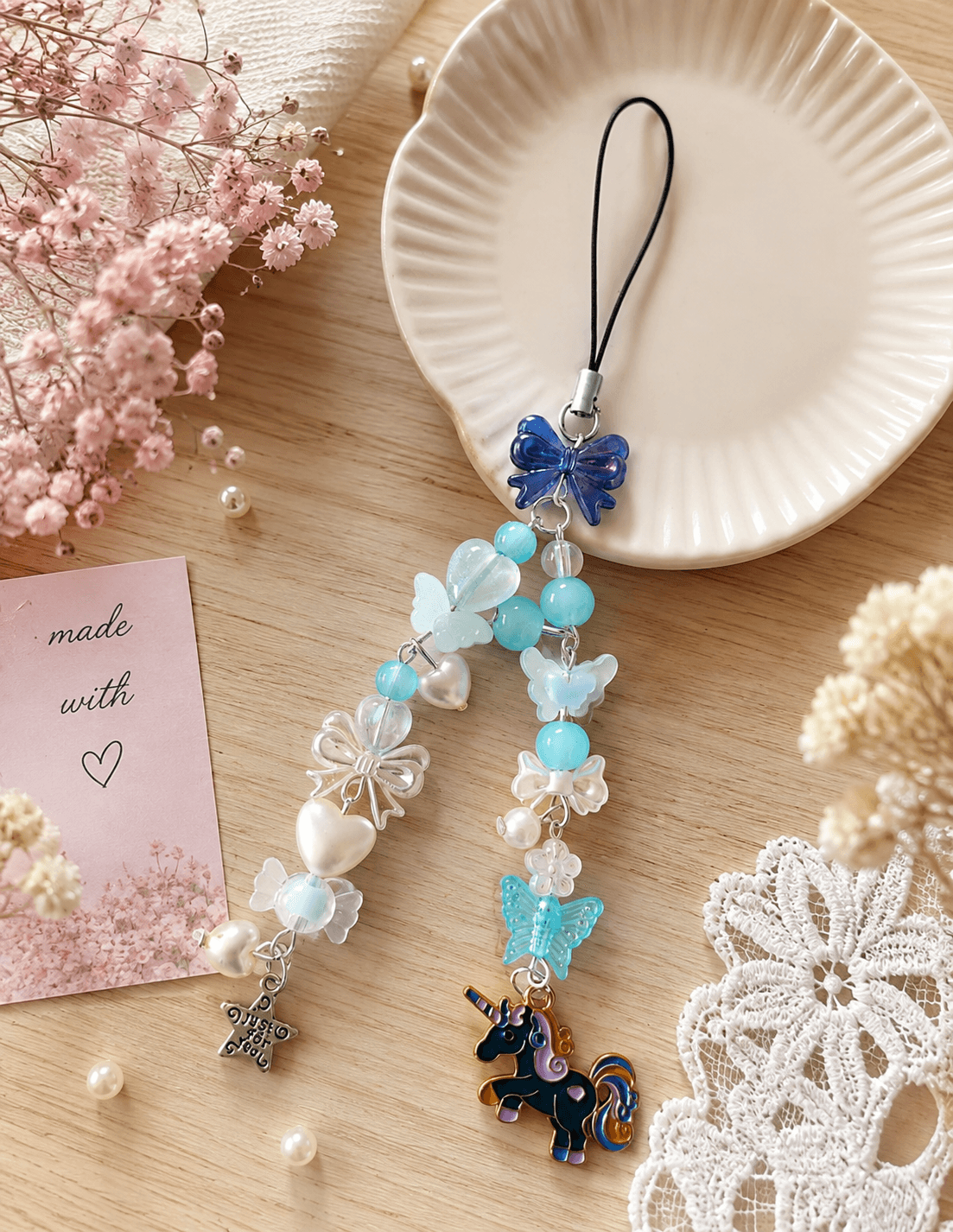Handmade aqua and white phone charm strap displayed with a dark blue bow, butterfly details, heart and flower beads, star charm and unicorn charm.